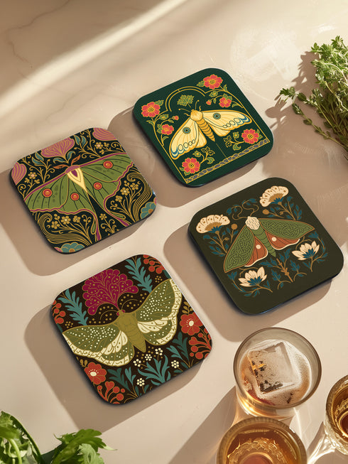 A set of four square hardboard coasters with smoothly rounded corners and a protective high-gloss finish. Each coaster features a unique, highly detailed Art Nouveau style illustration of a moth centered against a dark, botanical background. The intricate artwork uses a rich, moody color palette of deep forest greens, ochre, muted olive, crimson, and black, evoking a vintage, scholarly feel. These decorative drinkware accessories are designed for coffee table and bar cart use.